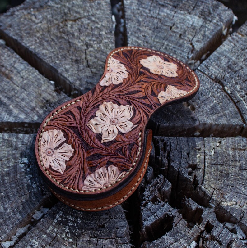 A brown leather pouch with a floral design. The pouch is decorated with a carved floral pattern in a light brown color. The coin purse is closed with a button .