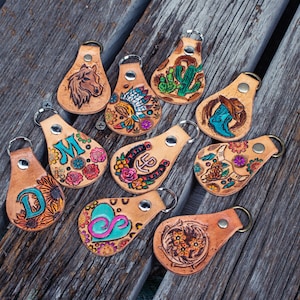 May include: Ten leather keyrings with colourful designs. The keyrings feature a variety of designs, including a horseshoe, a cactus, a boot, a sunflower, a heart, and a bull's head. The keyrings are all made of brown leather and have a metal ring for attaching keys.