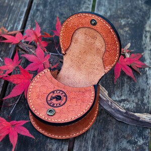 May include: A brown leather pouch with a flap closure and a decorative embossed design. The pouch is open and the inside is visible. The pouch is sitting on a wooden surface.