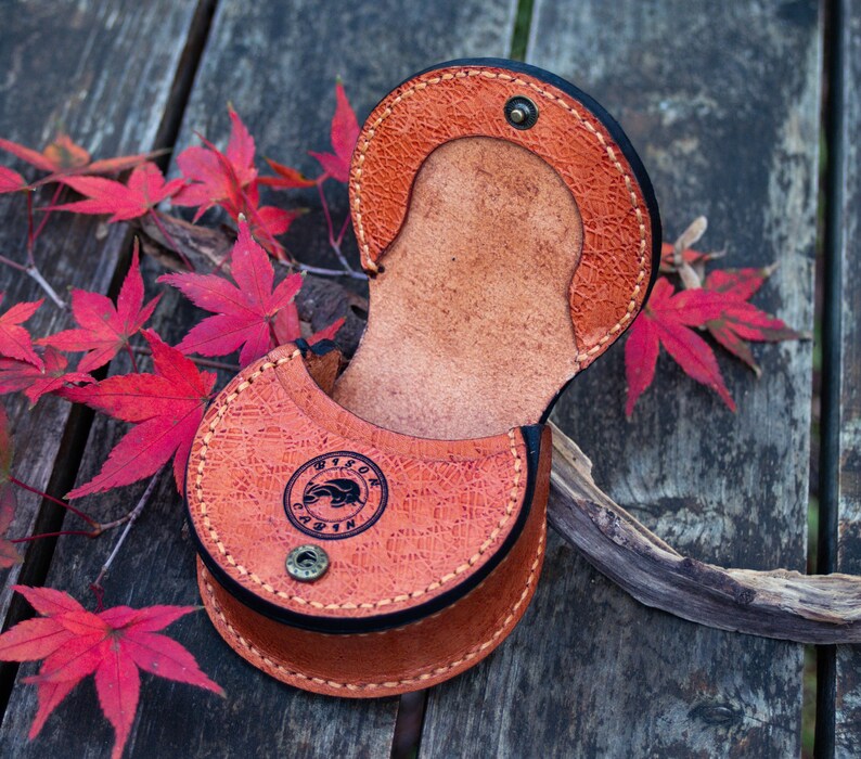 May include: A brown leather pouch with a flap closure and a decorative embossed design. The pouch is open and the inside is visible. The pouch is sitting on a wooden surface.