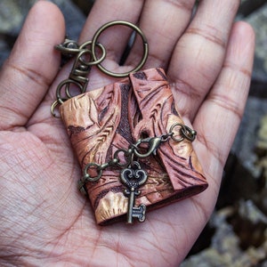May include: A small, brown leather journal with a key charm attached to a keyring. The journal has an intricate floral design embossed on the cover. The key charm is made of metal and has a vintage look.