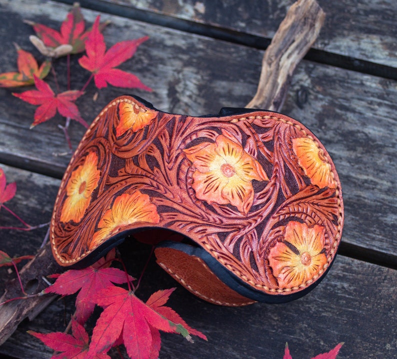 A leather coin wallet with a floral design. The wallet is brown with orange flowers and yellow color . The wallet is button closed and hand stitching