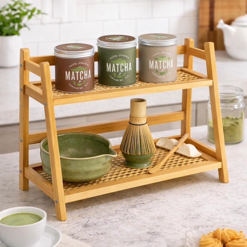 May include: A bamboo two-tiered shelf displays three matcha tea tins, a green ceramic bowl, a whisk, and a wooden spoon. The tins are brown, green, and beige, each labelled "Matcha Japanese Tea". A cup of matcha tea is in the foreground.