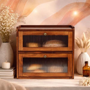 May include: A two-tiered wooden bread box with glass paneled doors. The box is made of dark brown wood and has a small brass knob on each door. The glass panels have a ribbed texture. The bread box is sitting on a white surface.
