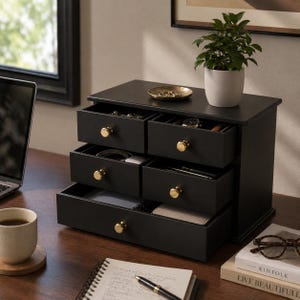 May include: A black wooden jewelry organizer with multiple drawers and gold-colored knobs. The top of the organizer has a small gold tray and a potted plant. The drawers are open, revealing various items. The organizer sits on a wooden desk.