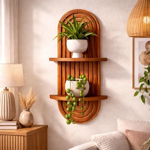 May include: A wooden wall shelf with two shelves and an arched top. The shelf is stained a warm brown and holds two white ceramic pots with green plants. The design includes vertical grooves and a semi-circular bottom.