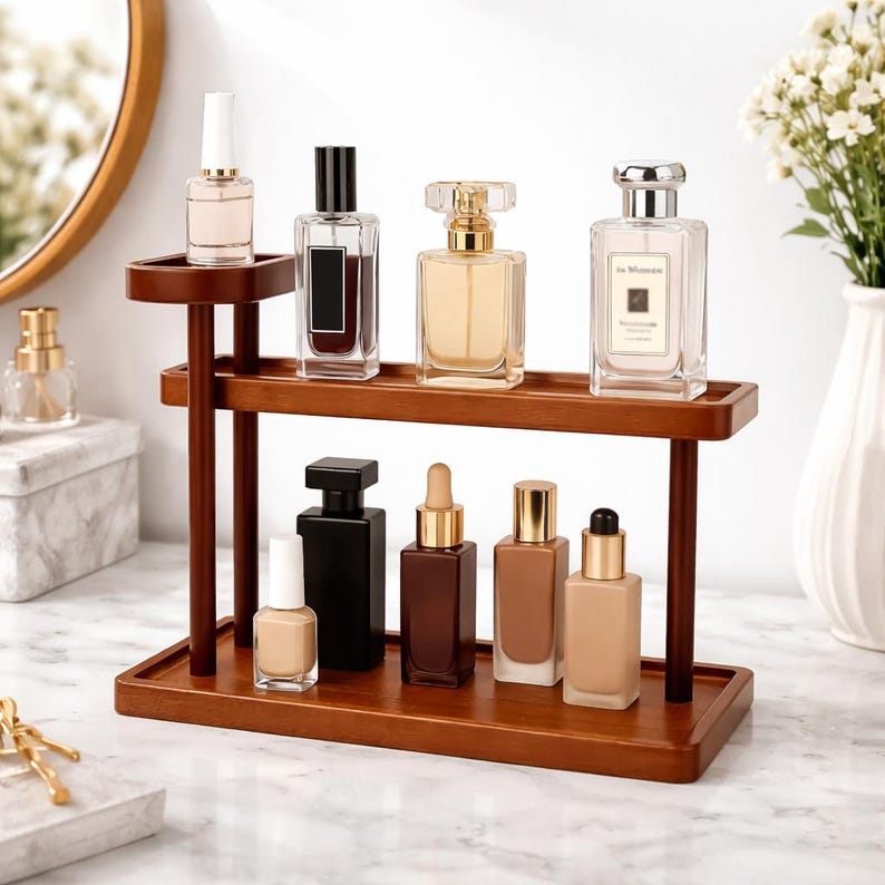 May include: A wooden display stand with two levels, showcasing perfume bottles and nail polish. The bottles are clear, with diverse shapes and lids. The stand is on a marble surface. A mirror and vase are visible in the background.
