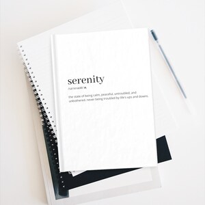 May include: A white notebook with the word "serenity" written in black text. The definition of the word is written below in smaller black text. The notebook is sitting on top of another notebook with a spiral binding.