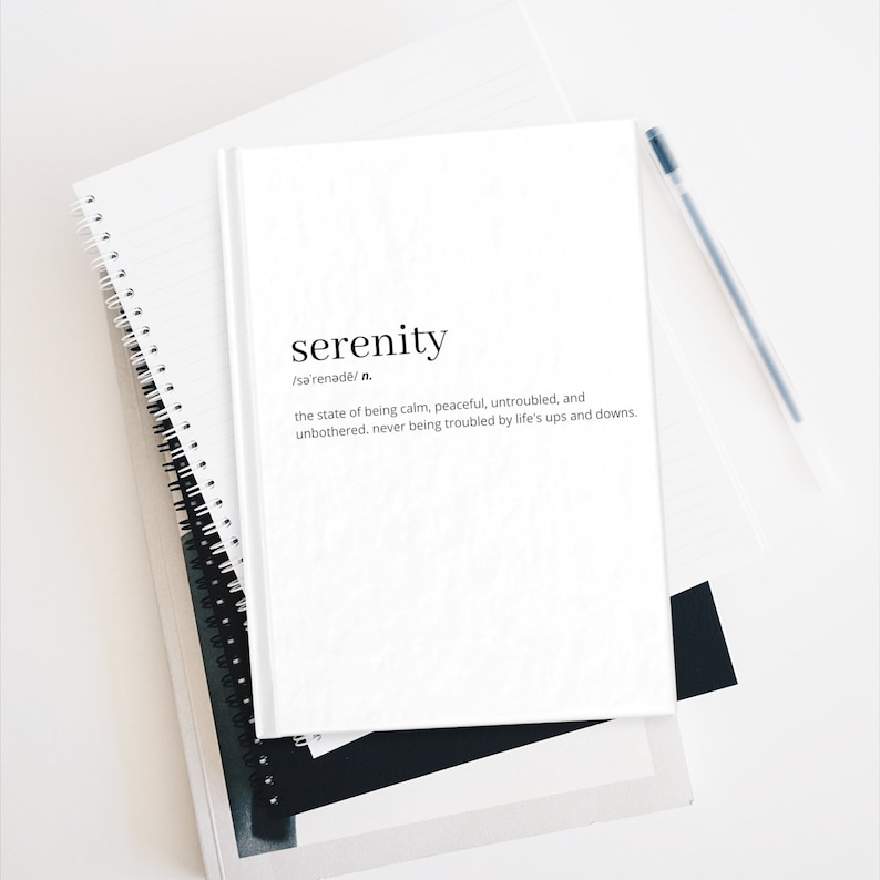 May include: A white notebook with the word "serenity" written in black text. The definition of the word is written below in smaller black text. The notebook is sitting on top of another notebook with a spiral binding.