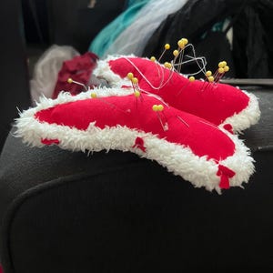 May include: A red butterfly-shaped fabric with white fur trim is pinned to a black surface with yellow pins.