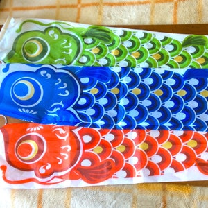 May include: Three colourful Japanese carp windsocks, also known as koinobori, in green, blue, and red. The windsocks are made of fabric and have a white background with a traditional Japanese design.