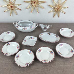 Könnte beinhalten: Ein weißes Porzellan-Tafelservice mit floralem Muster und rotem Rand. Das Set umfasst eine Terrine, eine Sauciere, Servierplatten und mehrere Teller. Dekorative Strohsterne hängen im Hintergrund.