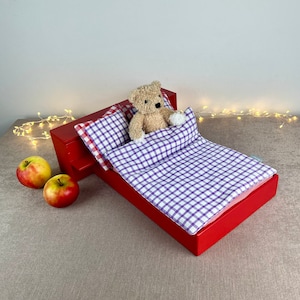 Può includere: Un letto di legno rosso per bambole con un set di biancheria da letto a quadri viola e bianchi. Un orsacchiotto è sdraiato sul letto. Due mele rosse sono sul pavimento davanti al letto.