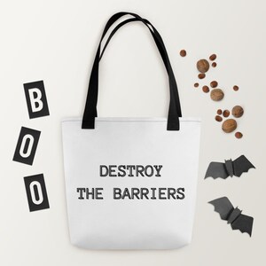 K&ouml;nnte beinhalten: Wei&szlig;e Einkaufstasche mit schwarzen Henkeln und dem Text "DESTROY THE BARRIERS" in Schwarz gedruckt. Die Tasche befindet sich auf einem wei&szlig;en Hintergrund mit schwarzen Buchstaben, die "BOO" buchstabieren, und zwei schwarzen Papierflederm&auml;usen.