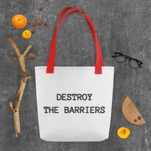 K&ouml;nnte beinhalten: Wei&szlig;e Einkaufstasche mit roten Henkeln und dem Text "DESTROY THE BARRIERS" in Schwarz gedruckt.