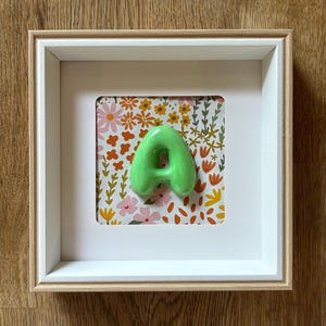 Può includere: Una lettera A tridimensionale, lucida e di colore verde lime, montata al centro di una cornice quadrata. Lo sfondo dietro la lettera presenta un motivo ripetitivo di piccoli fiori rosa, arancioni e gialli su una superficie bianca.