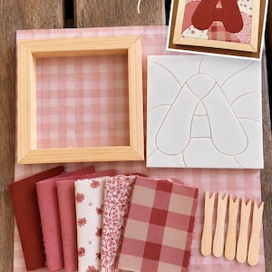Può includere: Un kit per il fai da te composto da una cornice quadrata in legno, una sagoma in plastica bianca con la lettera A in rilievo, cinque campioni di tessuto nei toni del rosso e del rosa con motivi floreali e a quadri, e cinque piccoli utensili in legno.