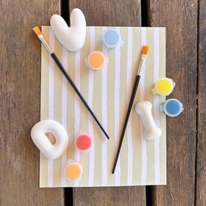 Può includere: Un kit artigianale con un oggetto a forma di cuore e un oggetto a forma di osso, un oggetto a forma di ciambella, pennelli e piccoli vasetti di vernice blu, arancione, gialla e rossa. Gli oggetti sono su carta a righe.