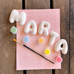Könnte beinhalten: Ein Set aus fünf weißen, abgerundeten, dreidimensionalen Buchstaben, die den Namen MARTA bilden und auf einer rosa Karte mit Gittermuster angeordnet sind. Das Set enthält einen dünnen Pinsel mit schwarzem Stiel und fünf kleine, runde Farbtöpfe in Grün, Blau, Orange, Gelb und Pink.
