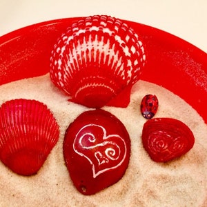 Könnte beinhalten: Eine Sammlung roter Muscheln und dekorativer Gegenstände, die auf Sand in einer roten Schüssel angeordnet sind. Die Muscheln umfassen eine große, gemusterte Muschel, eine Muschel mit Herzmuster und einen kleinen Marienkäfer.