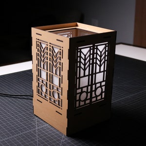 Könnte beinhalten: Eine braune Holzlampe mit geometrischem Design. Die Lampe hat einen weißen Papierschirm und ist beleuchtet.