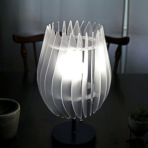 Puede incluir: Una lámpara de mesa blanca con una pantalla única en forma de flor geométrica. La pantalla está hecha de paneles translúcidos superpuestos que crean un aspecto delicado y aireado. La lámpara está sobre una base negra y está encendida, proyectando un brillo cálido.