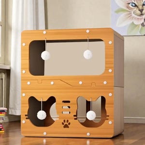 May include: A wooden cat house with a light brown finish. The house has two levels, with cat-shaped cutouts and hanging toy balls. The design includes a paw print and a large rectangular opening. White circular accents are placed around the edges.