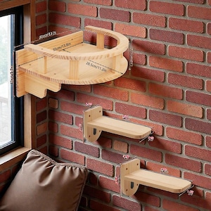 Puede incluir: Un conjunto de estantes y una cama curva para gatos de madera, montados en una pared de ladrillo. La cama para gatos mide 35 cm de ancho y 28,5 cm de profundidad. Los estantes son rectangulares y tienen soportes metálicos.