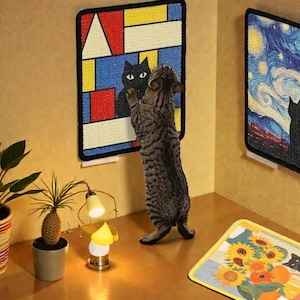 Könnte beinhalten: Eine getigerte Katze steht auf ihren Hinterbeinen und greift nach einem gerahmten Kunstwerk, das eine schwarze Katze darstellt. Das Kunstwerk ist ein farbenfrohes, abstraktes Design. Ein weiteres gerahmtes Stück zeigt eine Sternennachtszene mit einer Katzen-Silhouette. Eine kleine Lampe und Topfpflanzen sind ebenfalls zu sehen.