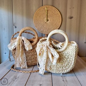 Op de afbeelding: Twee geweven strooien handtassen met ronde handvatten en crèmekleurige kanten sjaals die aan de handvatten zijn vastgebonden. Een ronde geweven placemat hangt aan de houten muur achter de tassen. De tassen staan op een houten oppervlak.