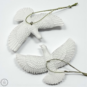 Könnte beinhalten: Zwei weiße Taubenornamente mit ausgebreiteten Flügeln, detailliert mit strukturierten Federn. Jede Taube hat eine goldfarbene Schnur zum Aufhängen. Die Ornamente sind vor einem weißen Hintergrund platziert.