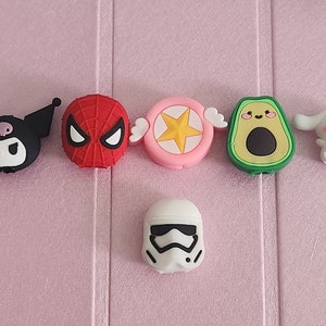 May include: Six colorful character phone charms, including a black and white Hello Kitty, a red Spiderman, a pink star with wings, a green avocado, a white Hello Kitty, and a white Stormtrooper.