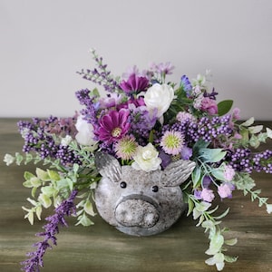 May include: A metal pig-shaped planter filled with an assortment of purple, white, and green artificial flowers. The planter has a distressed, galvanized finish, and the flowers create a full, vibrant arrangement.
