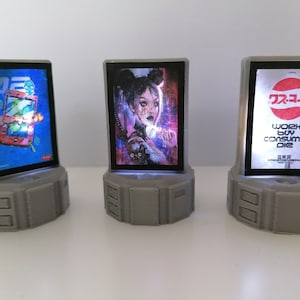 May include: Three miniature light-up display stands with different images. The first stand has a blue background with a cartoon image of a phone and a green alien. The second stand has a purple and pink background with a cartoon image of a woman with a black cat. The third stand has a white background with a red and white logo and the text "work buy consume die".