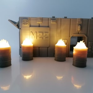 May include: Four grey metal barrels with flames on top, in front of a grey building with the letters "M22" on the side.