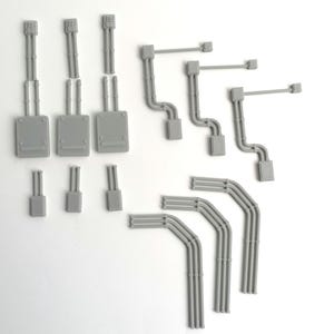 May include: A selection of grey plastic model components, including rectangular boxes, vertical tubes, and curved pipes. The parts are arranged on a white surface, possibly for a model or diorama.