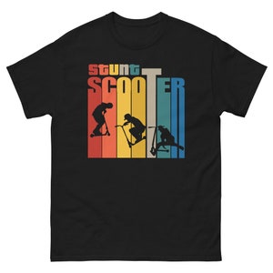 Op de afbeelding: Zwart T-shirt met een retro-grafiek van drie personen die op scooters rijden. De grafiek heeft een regenboogkleurenschema en de tekst "Stunt Scooter" is boven de grafiek gedrukt.