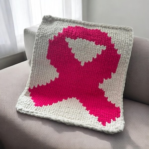 May include: A pink ribbon crocheted into a square throw pillow. The pillow is made with a chunky yarn and has a white background.