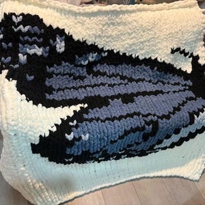 May include: A chunky knit blanket featuring a large, stylized whale graphic. The design is created with thick, soft yarn in shades of cream, black, and various tones of blue, forming a distinct contrast against the cream-colored background.