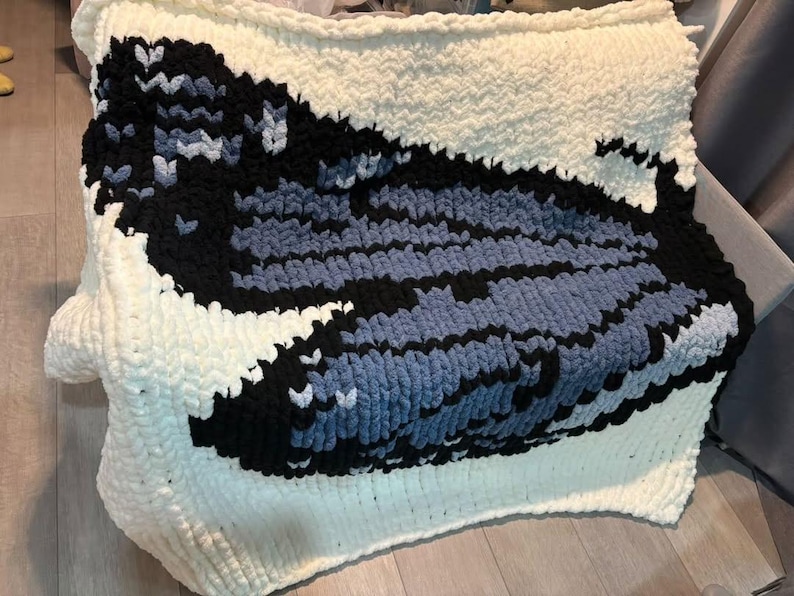 May include: A chunky knit blanket featuring a large, stylized whale graphic. The design is created with thick, soft yarn in shades of cream, black, and various tones of blue, forming a distinct contrast against the cream-colored background.