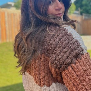 May include: A color-blocked sweater featuring a thick, textured, chunky knit pattern in shades of cream, light brown, and dark brown. The garment has a soft, plush appearance with a distinct ribbed construction throughout the body and sleeves.