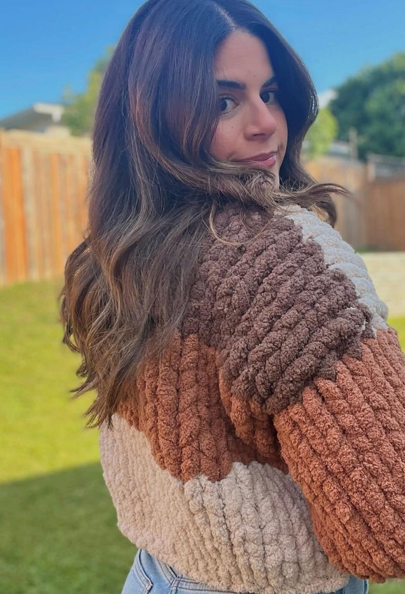 May include: A color-blocked sweater featuring a thick, textured, chunky knit pattern in shades of cream, light brown, and dark brown. The garment has a soft, plush appearance with a distinct ribbed construction throughout the body and sleeves.