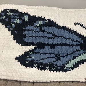 May include: A rectangular chunky knit blanket featuring a large butterfly wing pattern. The design is composed of thick white, navy blue, medium blue, and light mint green yarn, creating a bold graphic contrast across the entire surface.