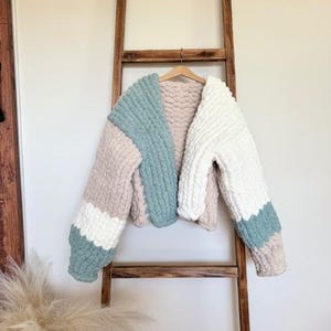 May include: A chunky knit cardigan featuring a color-blocked design in sage green, cream, and beige. The garment is crafted from thick, soft yarn with a visible looped texture, long sleeves, and an open front, displayed on a wooden hanger.