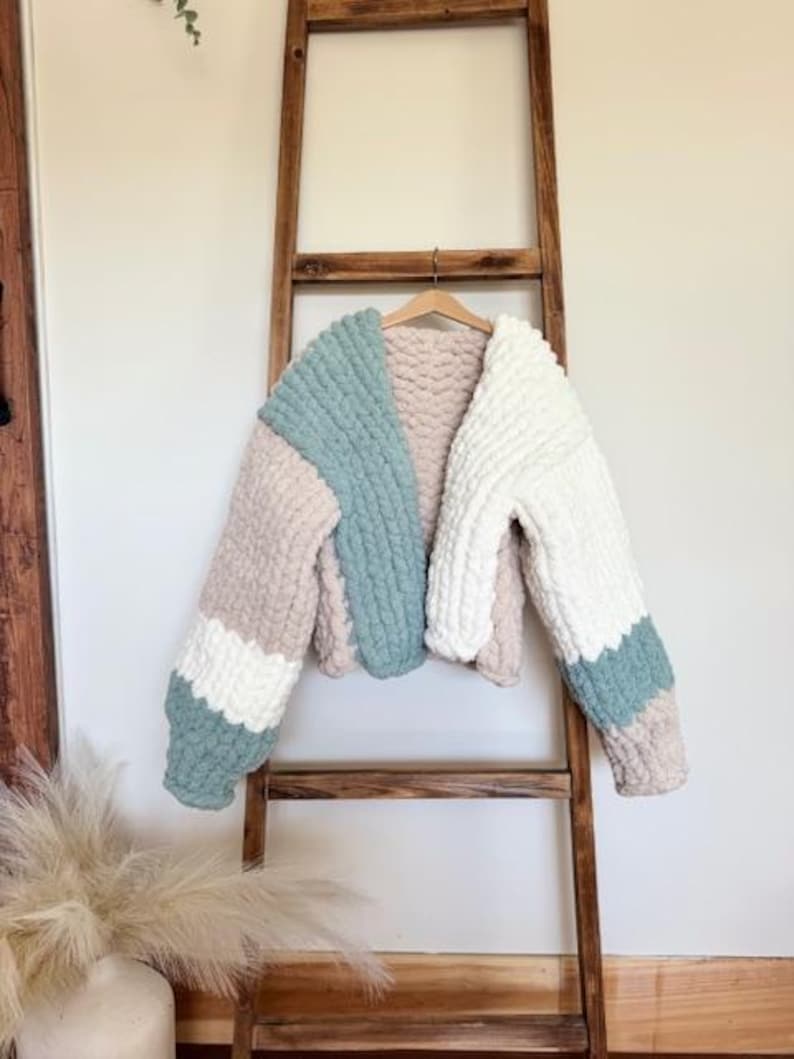 May include: A chunky knit cardigan featuring a color-blocked design in sage green, cream, and beige. The garment is crafted from thick, soft yarn with a visible looped texture, long sleeves, and an open front, displayed on a wooden hanger.