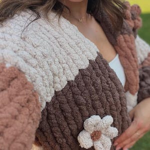 May include: A chunky knit cardigan featuring a color-blocked pattern in cream, dark brown, and light brown. The garment is crafted from thick, soft yarn and includes a prominent, textured flower appliqu&eacute; on the front panel with a brown center.
