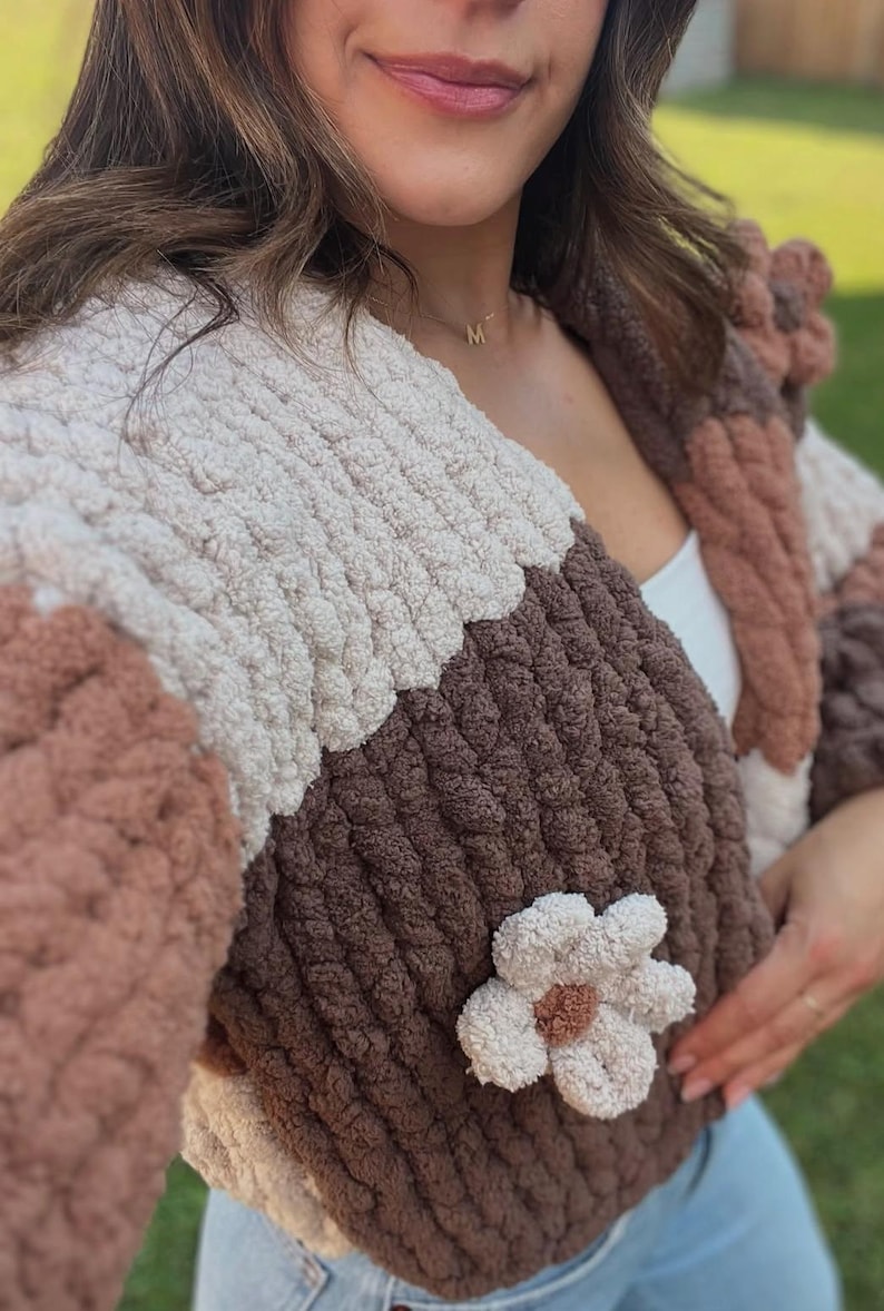 May include: A chunky knit cardigan featuring a color-blocked pattern in cream, dark brown, and light brown. The garment is crafted from thick, soft yarn and includes a prominent, textured flower appliqu&eacute; on the front panel with a brown center.