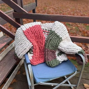 May include: A chunky knit blanket featuring a color-blocked pattern of cream, coral pink, olive green, and dark brown yarn. The throw is crafted from thick, soft, chenille-style yarn with a visible looped texture, draped over a blue folding chair.