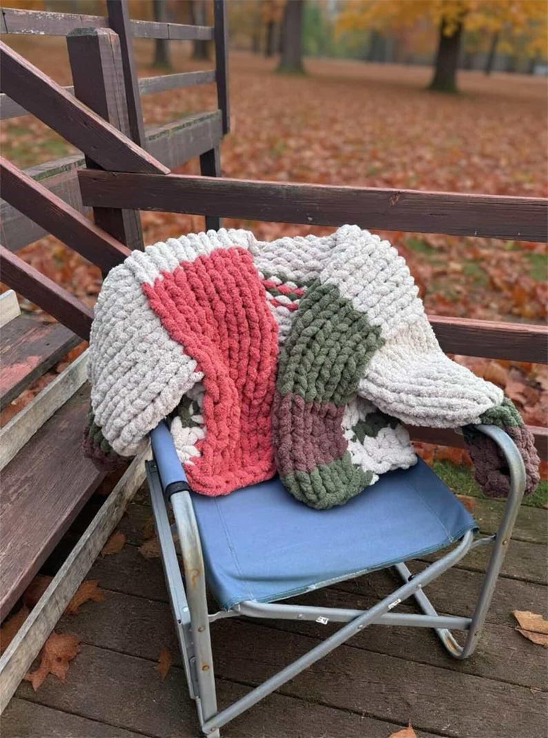 May include: A chunky knit blanket featuring a color-blocked pattern of cream, coral pink, olive green, and dark brown yarn. The throw is crafted from thick, soft, chenille-style yarn with a visible looped texture, draped over a blue folding chair.