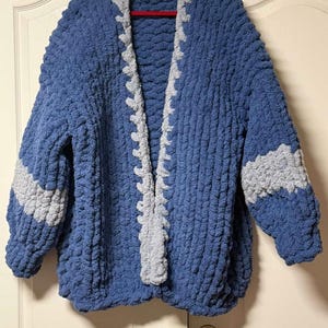 May include: A chunky knit cardigan made from thick blue yarn with a contrasting light gray trim along the open front edges. The sleeves feature a wide horizontal light gray stripe, and the entire garment has a textured, looped appearance.